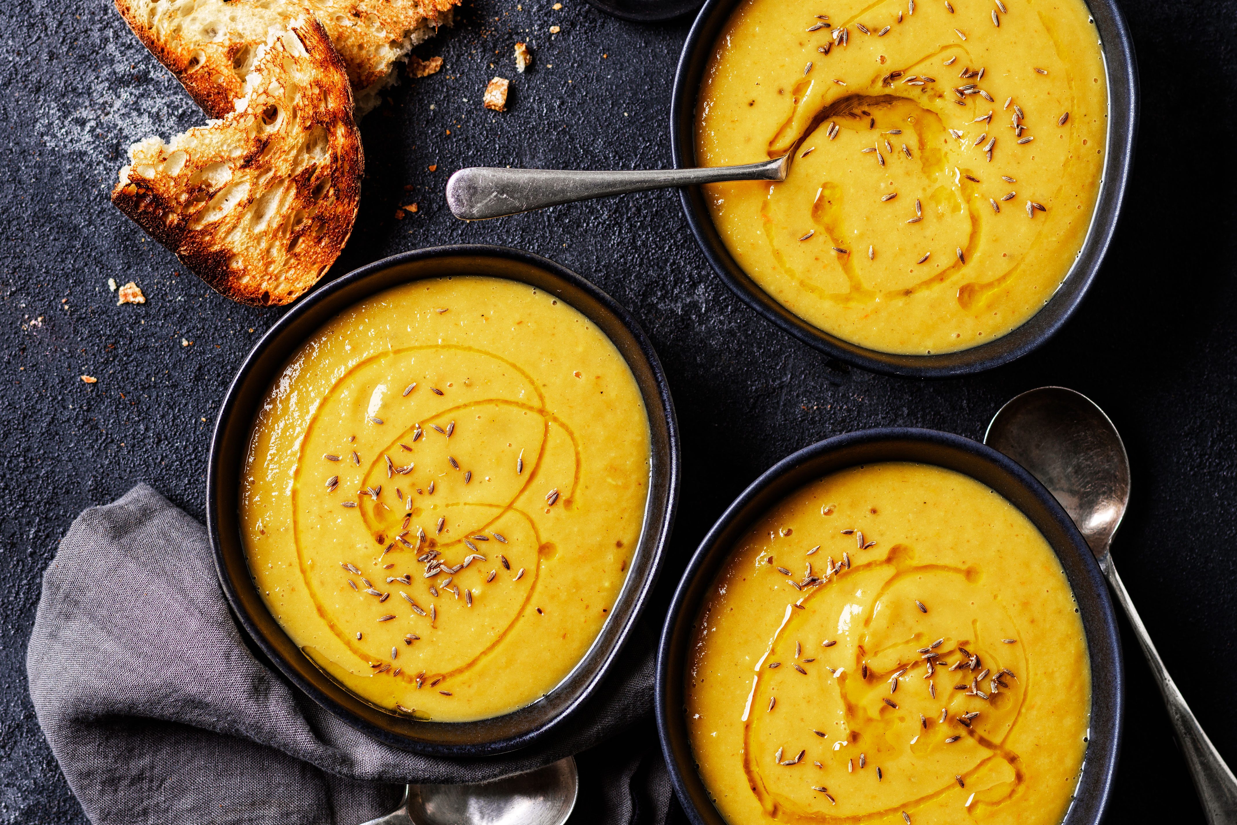 Three bowls of parsnip soup topped with oil and seeds, next to slices of bread