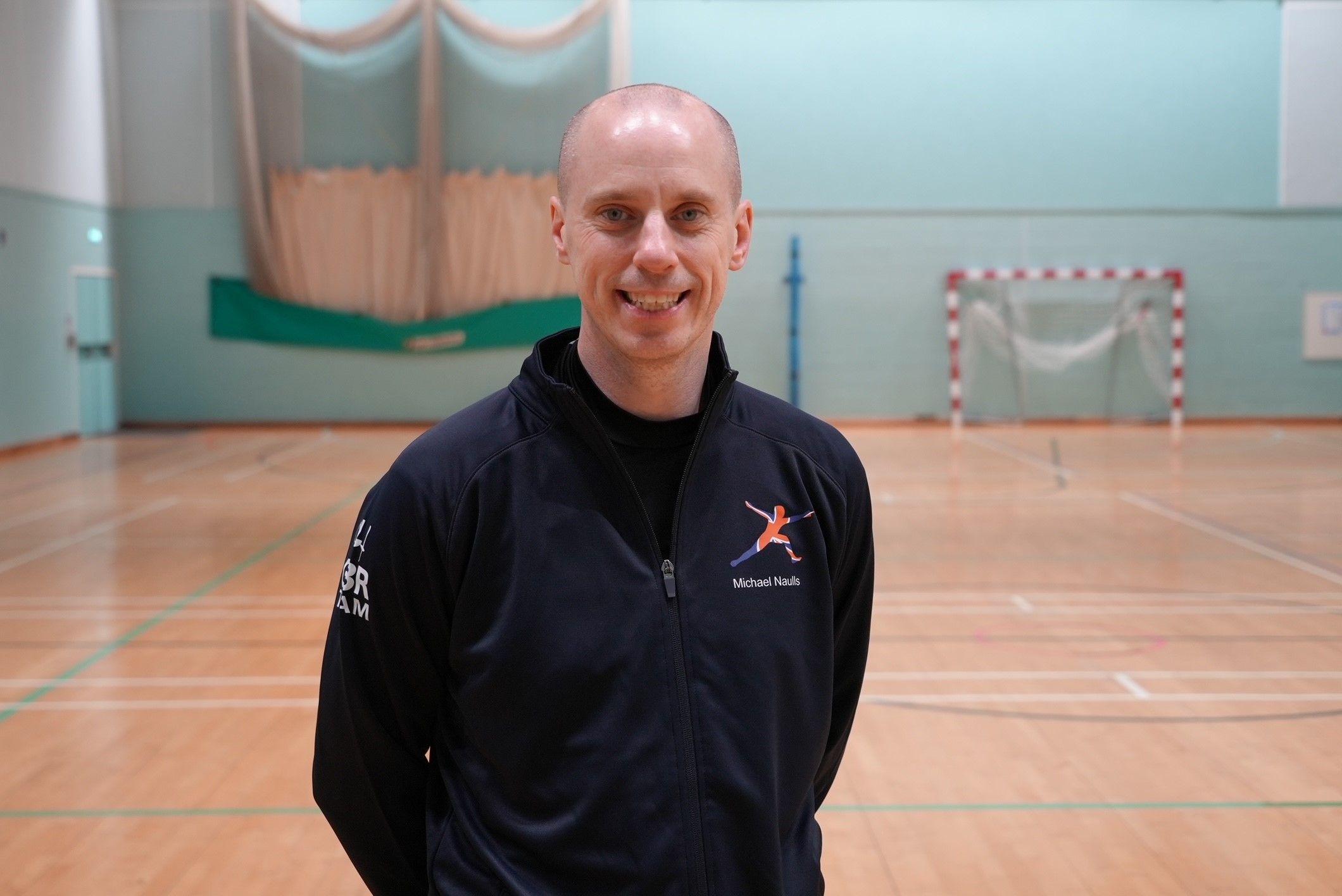 Michael Naulls in his navy blue British fencing training kit smiles for the camera. He's standing in a sports hall in Norfolk where he trains.