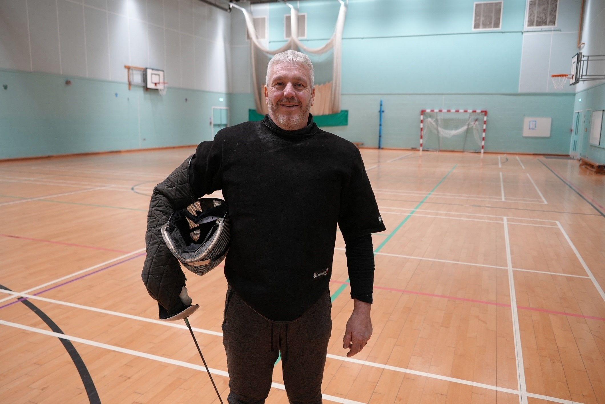 Coach Paul standing in a sports hall with a weapon in his right hand and fencing mask under his arm